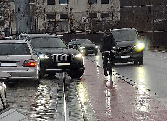 SUV steht beim Ausfahren aus Parkbucht halb auf rotem Fahrrad-Schutzstreifen, auf dem eng ein Radfahrer vorbeifährt, der ausweichen muss