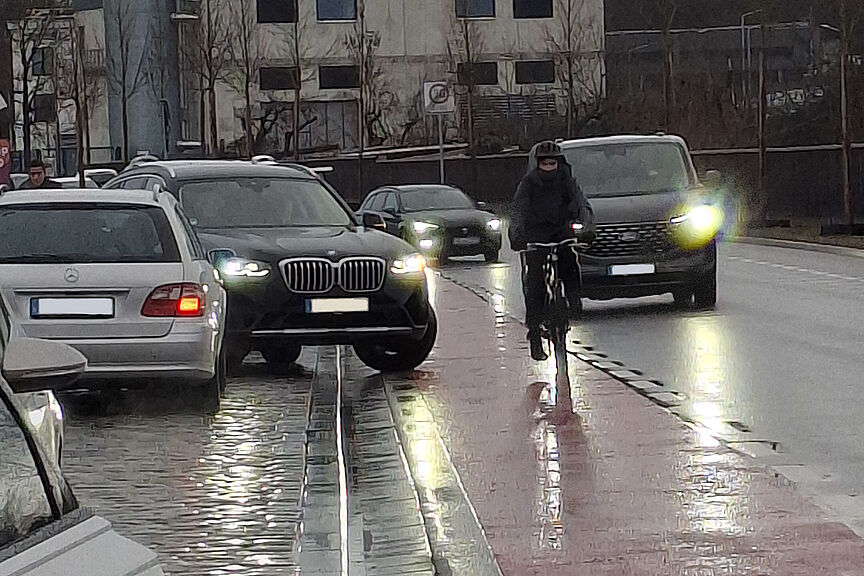 SUV steht beim Ausfahren aus Parkbucht halb auf rotem Fahrrad-Schutzstreifen, auf dem eng ein Radfahrer vorbeifährt, der ausweichen muss
