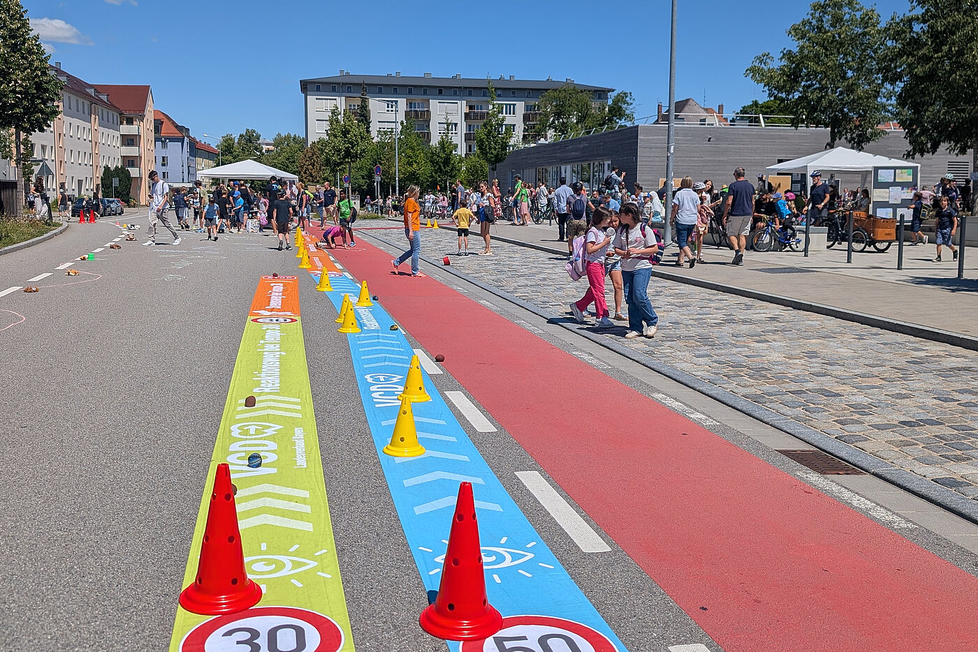 Banner auf der Straße verdeutlichen Anhalteweg bei Tempo 30 und Tempo 50. Dahinter unterhalten sich viele Menschen auf der Straße vor der Kreuzschule