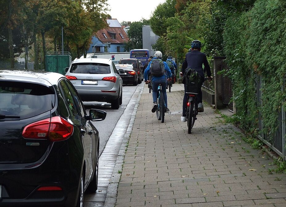 Auf dem Gehweg fahrende Schüler Radfahrende Schüler/-innen die auf dem Gehweg neben parkenden Fahrzeugen auf der Fahrbahn fahren